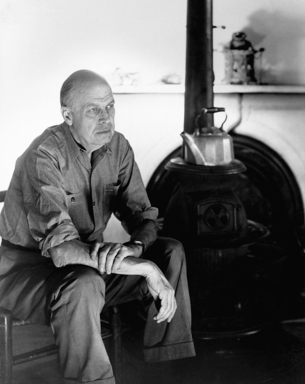 A b&w image; elderly Hopper sits, mostly bald, and turns slightly from the camera to his left, in front of an old fashioned stove and kettle; he grasps one arm with the other hand and wears a simple button down shirt and pants; wears a serious expression