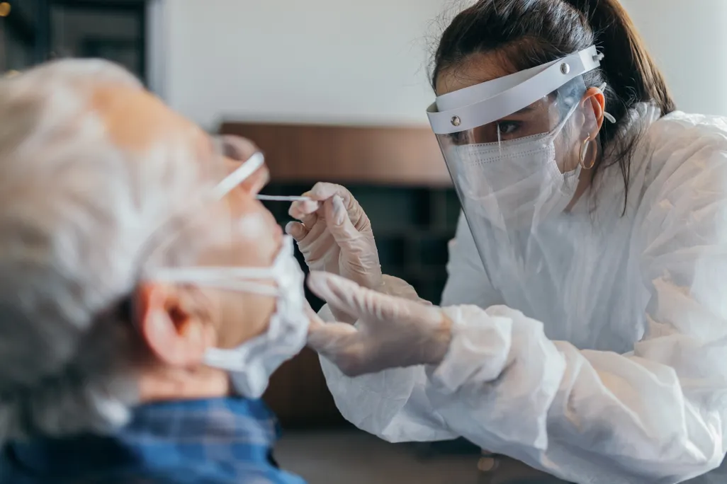 A female doctor in a mask and visor gives a nasal swab to a male patient.