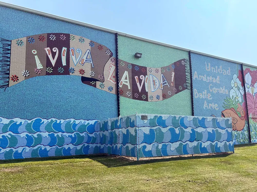 Turquoise mural covering the entire side of a building, with a pattern of waves along the bottom, and a brown textile with the Spanish words Viva la Vida! in the center.