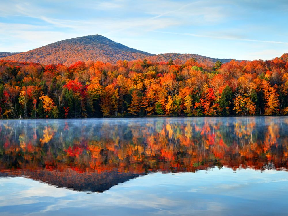 A lake, with a distant sloping hill and brilliant and orange red trees reflected perfectly in the still water. Overhead, a blue sky with wispy white clouds