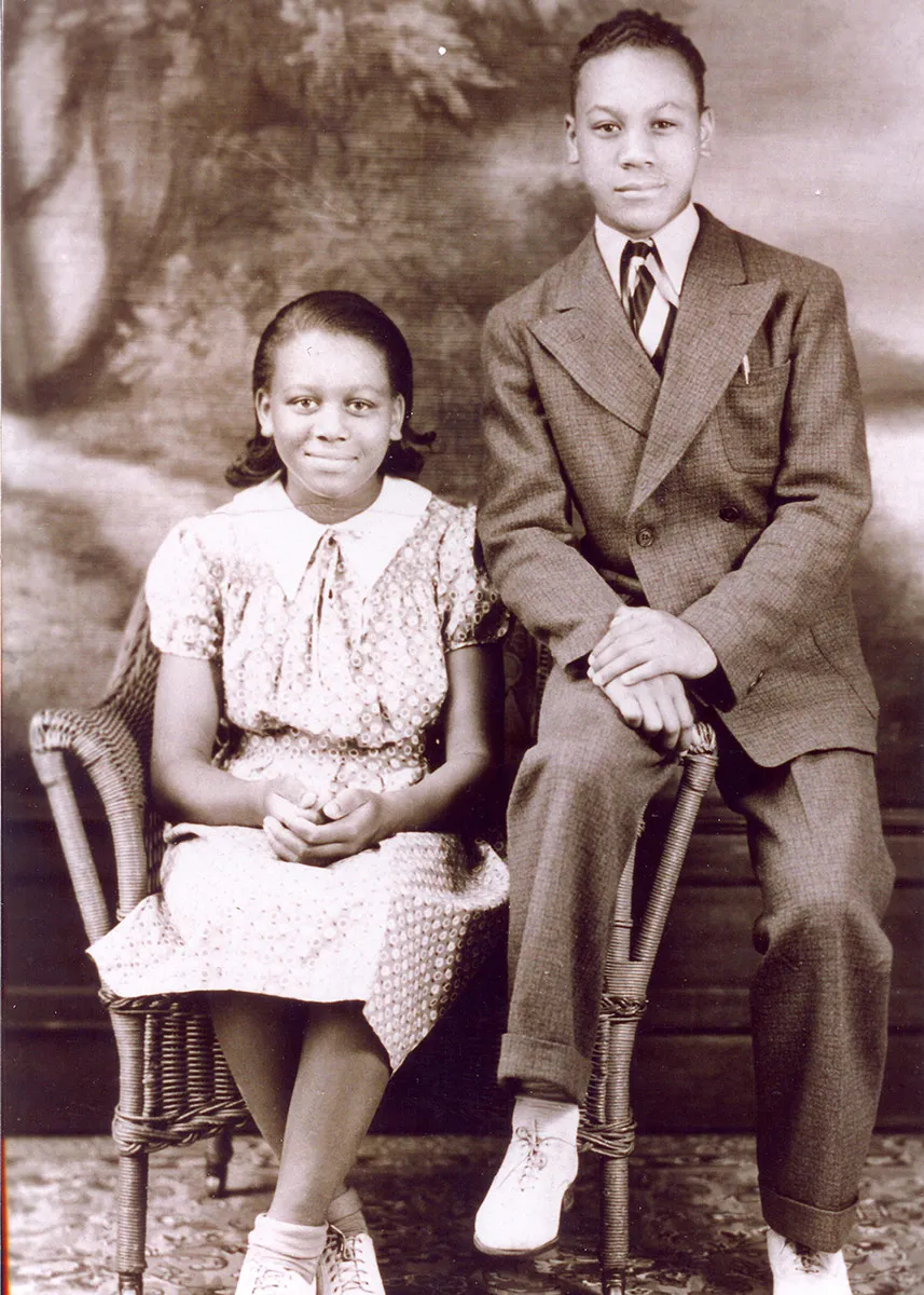 Studio portrait of a a girl and teen boy, posing on a wicker chair and wearing a dress and suit. Old sepia-toned photograph.