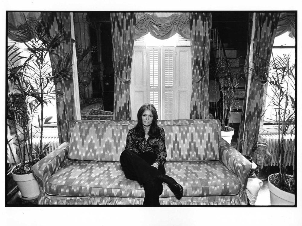 A black and white image of Steinem, a white woman in a patterned button down and black pants, sitting on a patterned couch in front of house plants and tall windows with curtains