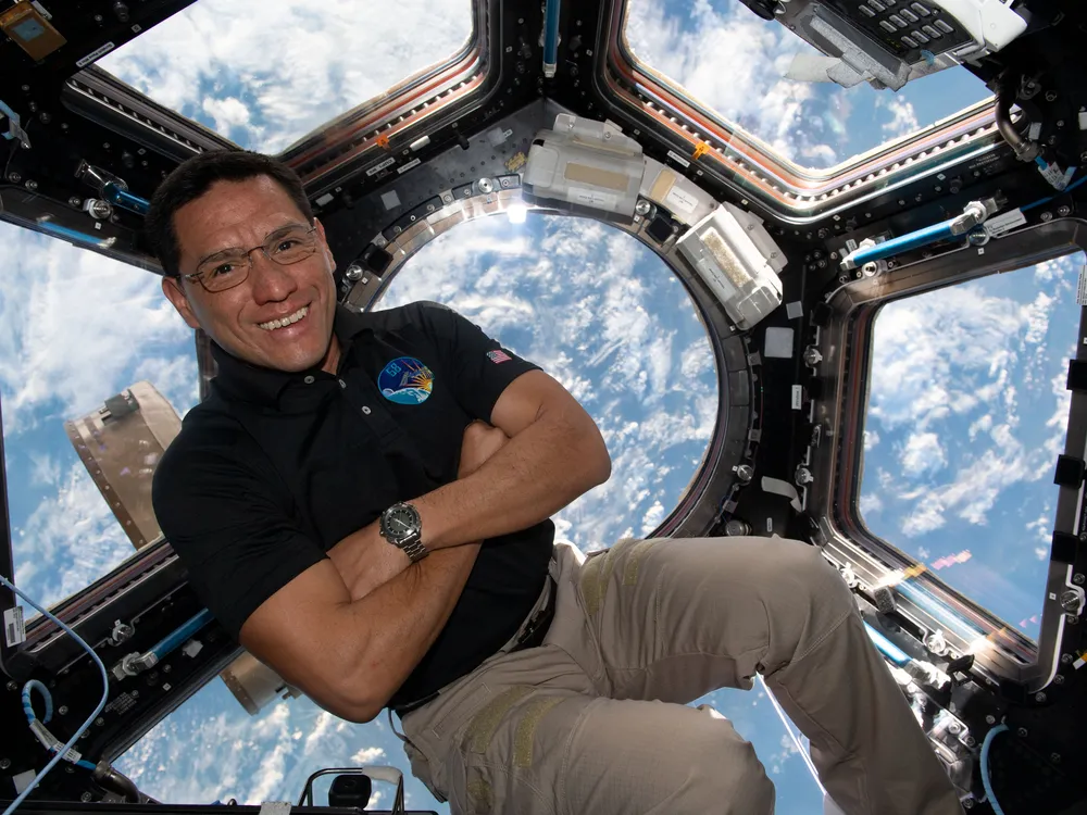 Frank Rubio floats with his arms crossed on the International Space Station with a view of Earth out a window behind him