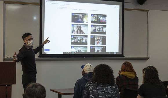 A man points to a video screen in a classroom with students seated in the foreground.