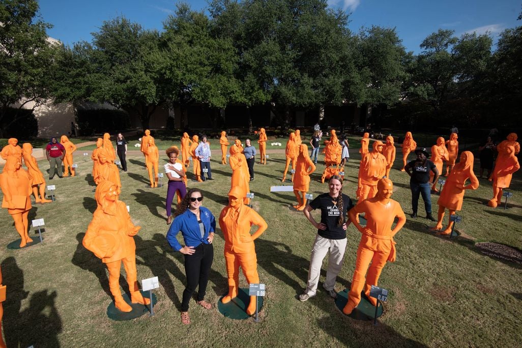 A field with life size orange plastic statues of women and two humans posing with them