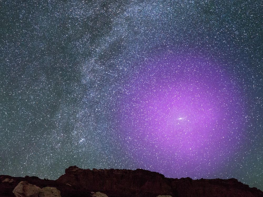 A view of rocks in foreground with an expansive, glittering night sky behind -- many stars and the curve of the Milky Way appear; in the lower righthand section of sky, an illustrated large circle of purple light indicates where Andromeda's halo would be