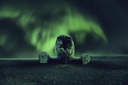 The northern lights cast an eerie glow upon an abandoned, wrecked airplane.

