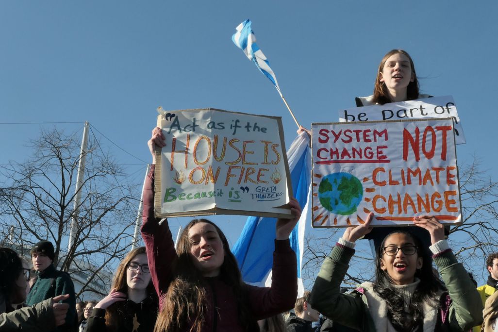 People gathered for a climate protest. There are several young people in the foreground holding signs that say: "Act as if the house is on fire because it is" and "System change not climate change."