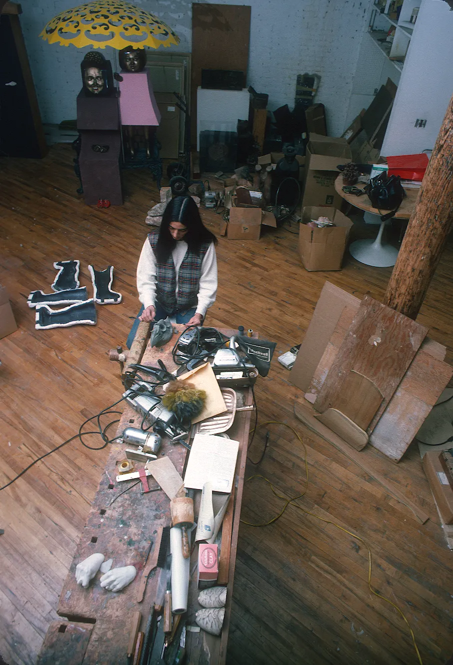 An artist in hr studio carving a clay mask and surrounded by supplies, molds, and a large sculpture. 