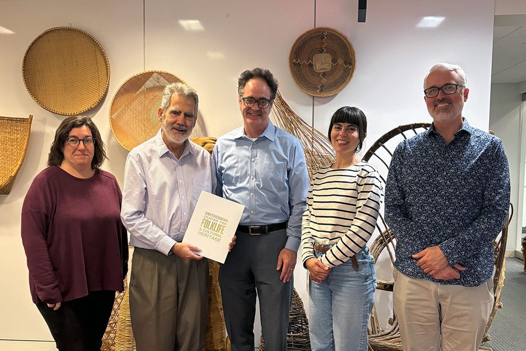 Five people pose smiling, lined up in a room with woven baskets hanging on the wall behind them. One man holds a document with the Smithsonian Center for Folklife and Cultural Heritage logo on the cover.
