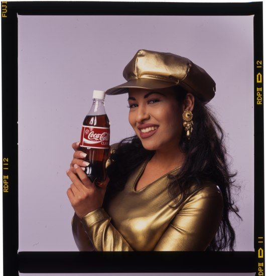 Selena in a gold cap holding a Coca-Cola bottle