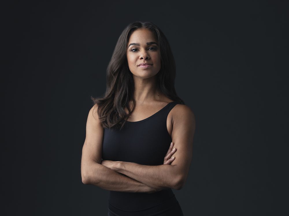 A woman stands with her arms crossed looking intently at the camera. She appears in front of a black background wearing a black leotard.