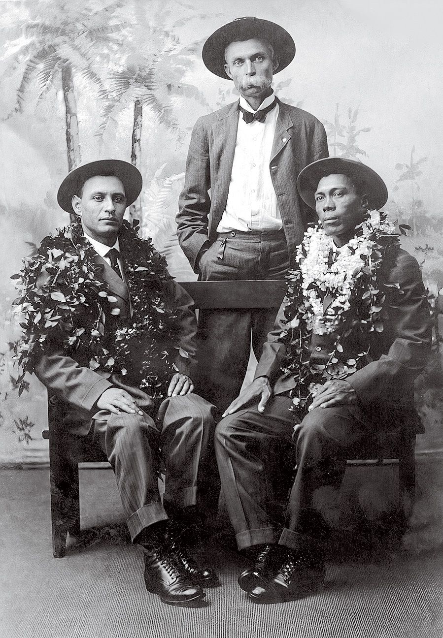 dva muži sedí s jedním mužem stojícím za portrétem