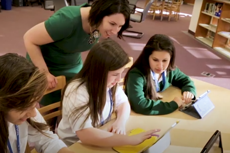 Students are working on their laptops and tablets while a teacher looks over their shoulders