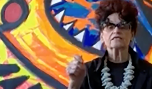 Detail of color screenshot of Judith Bernstein in front of one of her paintings wearing a black blouse and light-colored, chunky necklace holding her hand up in a fist as if making a point in conversation.