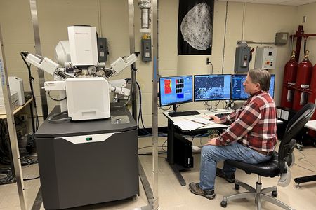 A man sits in front of a large, white machine and a row of computer screens in a lab setting.