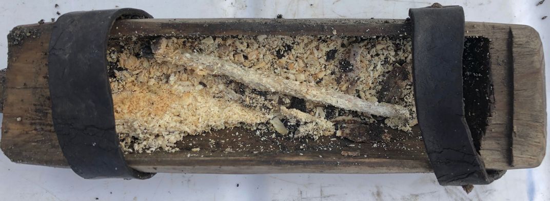 An interior view of the rectangular box, which is full of what resembles a long thin tapered candle, surrounded by crumbled bits of yellowed beeswax
