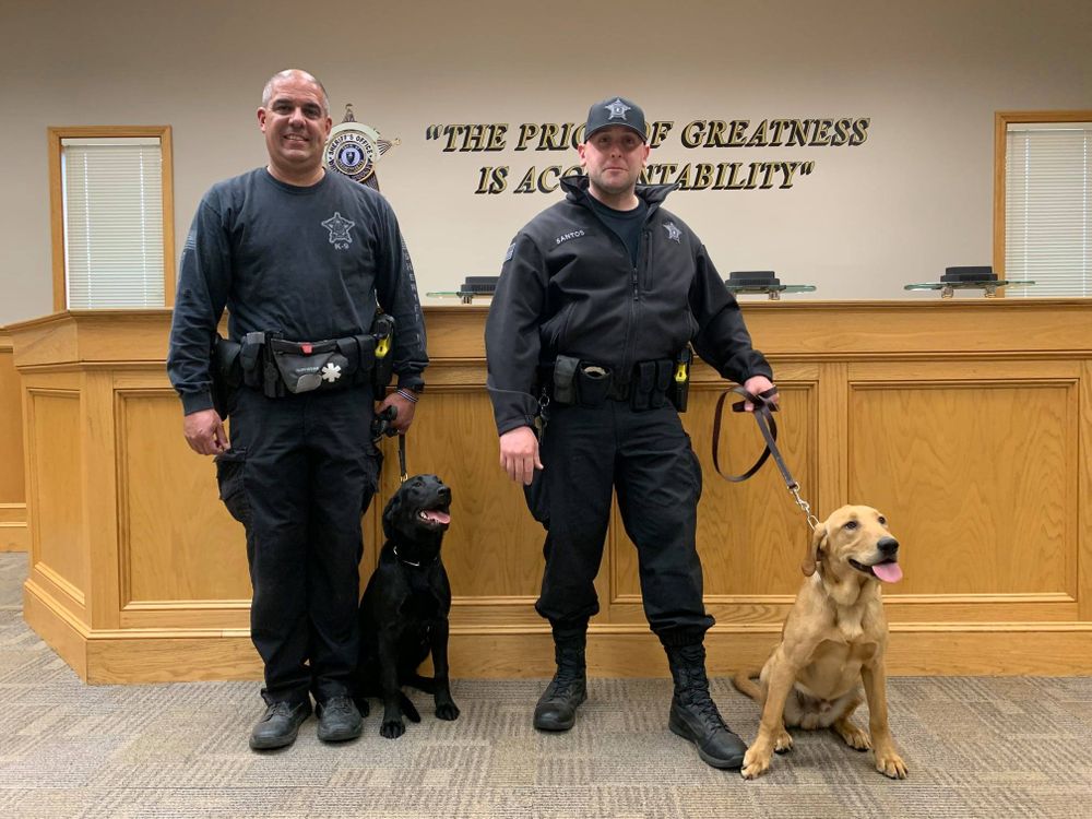Officers Paul Douglas (left) and Theodore Santos (right) stand with their newest Covid-19 K9 unit: a female black lab named Huntah (left) and a male golden lab-retriever mix, Duke (right).