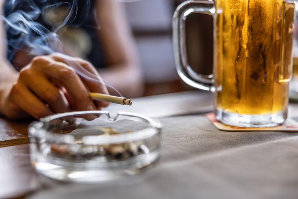 hand holding a lit cigarette near an ash tray and pint of beer