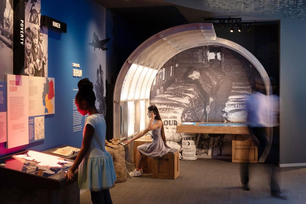 Two young people sit and stand in front of exhibits, reading the text; the blue walls are richly decorated with photographs, maps and information about post-war Europe
