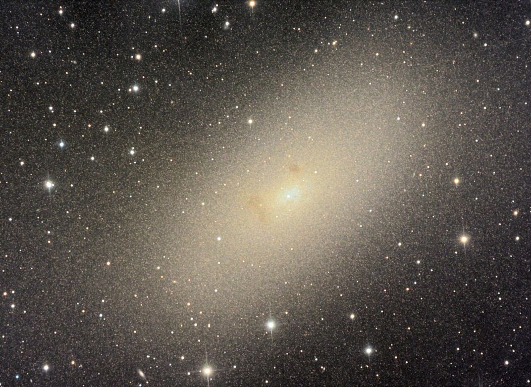 a yellow elliptical galaxy appears as a fuzzy blur in front of other galaxies and stars in the distance