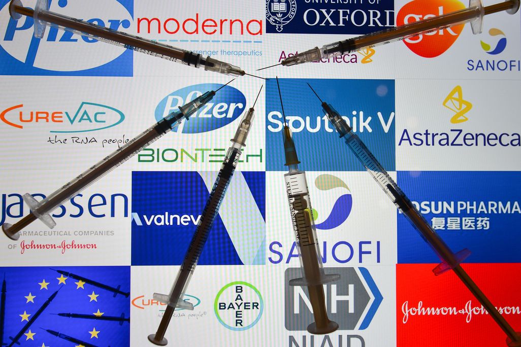 An illustrative image of medical syringes in front of the logos of various companies working on the Covid-19 vaccine, displayed on a screen. 