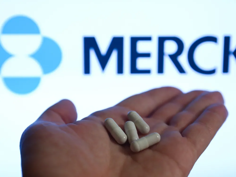 Image of left hand holding four white capsules against a Merck-branded backdrop