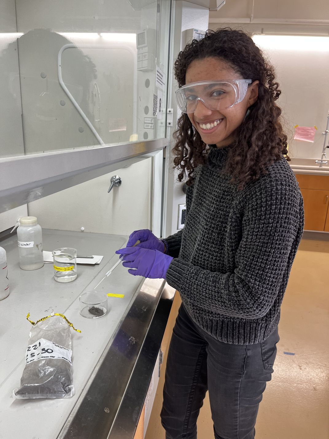 A young woman wearing goggles and purple gloves stands in a lab, preparing a sample on a table with a fume hood