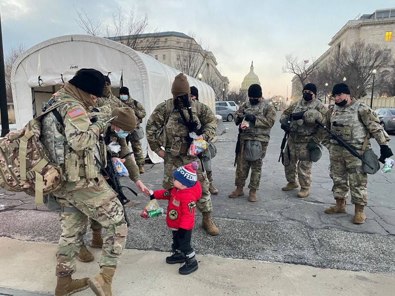A small child wearing a red coat decorated with insignia hands a bag of snacks to a group of uniformed members of the National Guard. The U.S. Capitol is visible in the background.