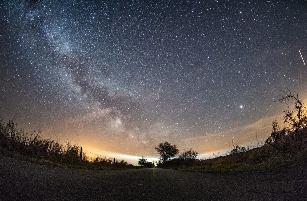 starry sky with the milky way visible and a treeline at the bottom, with a few streaks of meteors