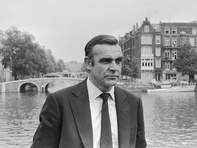 Image is black and white showing a man in a suit standing in front of a river