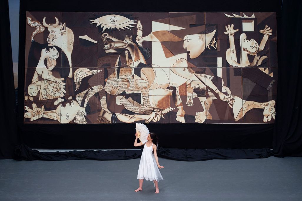 A person in a white flowy dress and holding a white fan dances in front of the enormous chocolate replica of Picasso's Guernica