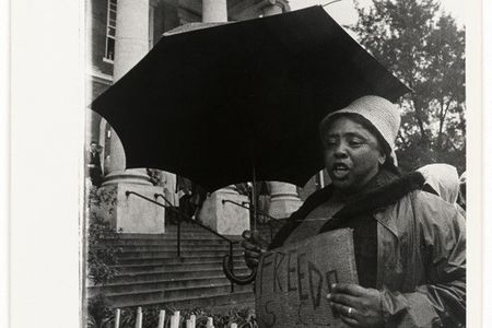 A black-and-white photograph of Fannie Lou Hamer carrying a sign and holding an umbrella over her head.