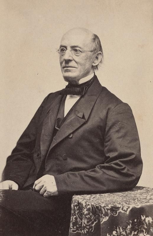A sepia-toned image of an elderly man, with a bald crown and glasses, in a suit, seated with his hand resting on a small table and in front of a blank white background