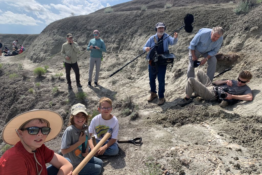 Three kids sitting with shovels on a hillside with a documentary crew in the background