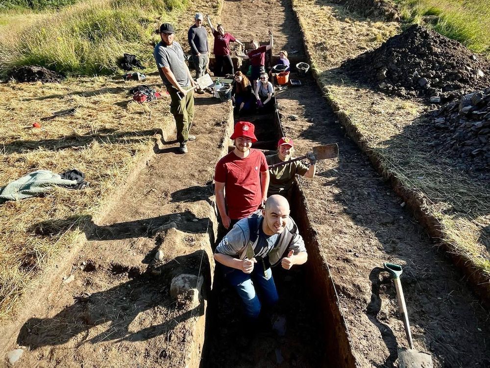 A group of people in a shallow ditch pose for a photo while digging