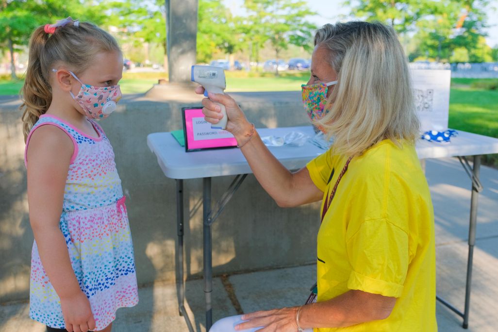 A young girl wearing a colorful cloth mask (left) gets her temperature read by a woman at the Childhood Development and Learning Lab at Central Michigan University