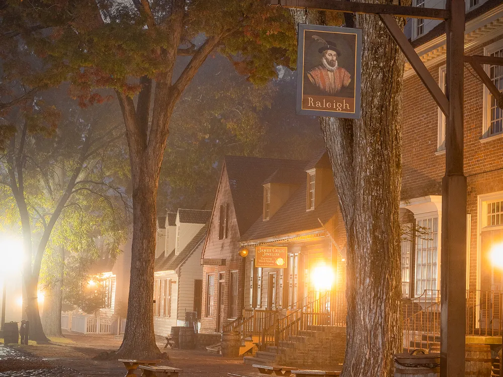 Colonial Williamsburg in autumn at dusk, with historic storefronts illuminated by warm light through a soft fog. Large trees with fall foliage line the street, and a hanging Raleigh sign adds to the scene’s colonial charm.