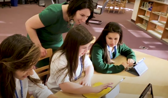 Students are working on their laptops and tablets while a teacher looks over their shoulders