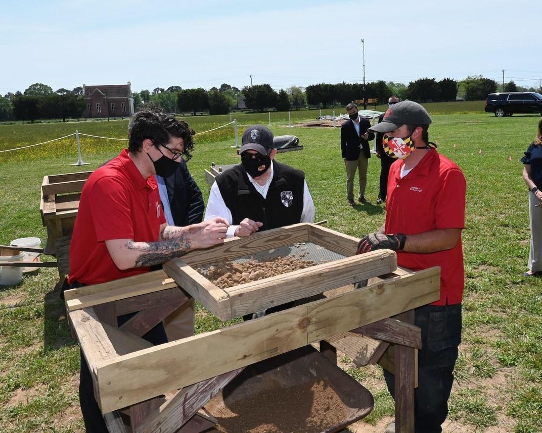 A group of people, some in red polo shirts and all wearing masks, stand outside in a field around a trough of dirt, examining an artifact