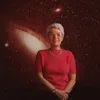 Vera Rubin stands against a backdrop of a star-filled galaxy with her hands loosely clasped in front of her.