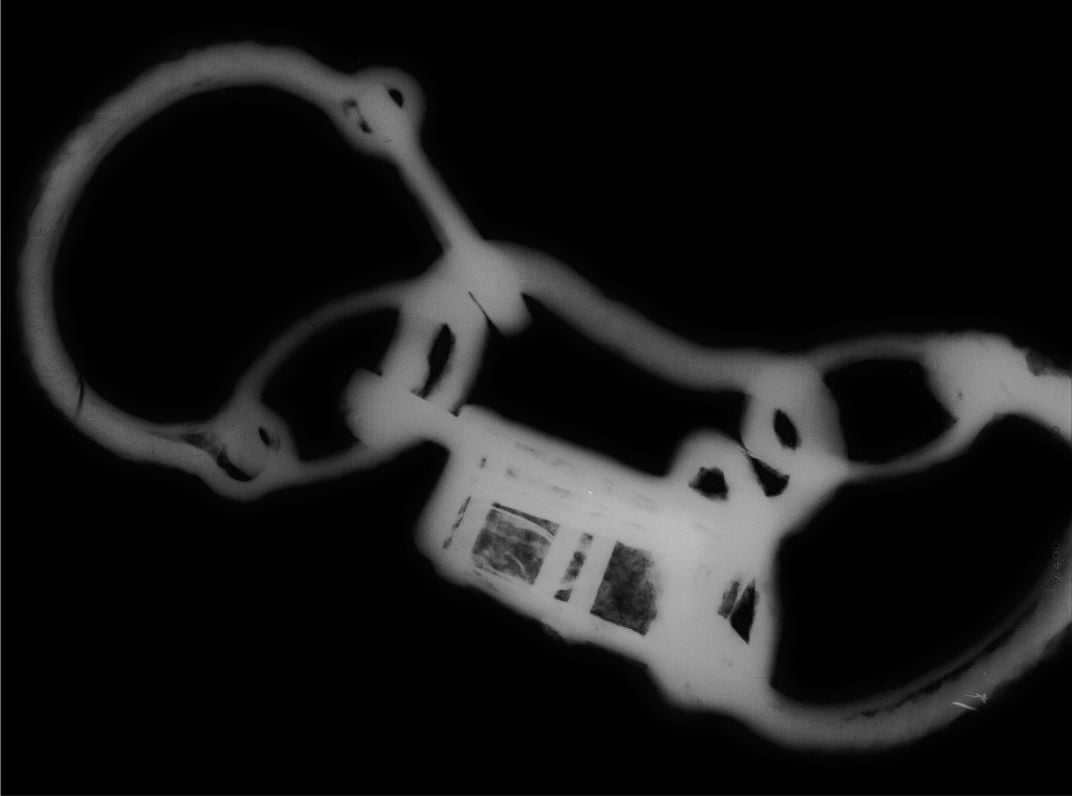 A black and white x ray image depicting the same set of shackles, with the small complicated internal mechanisms of the padlock, center, thrown into relief