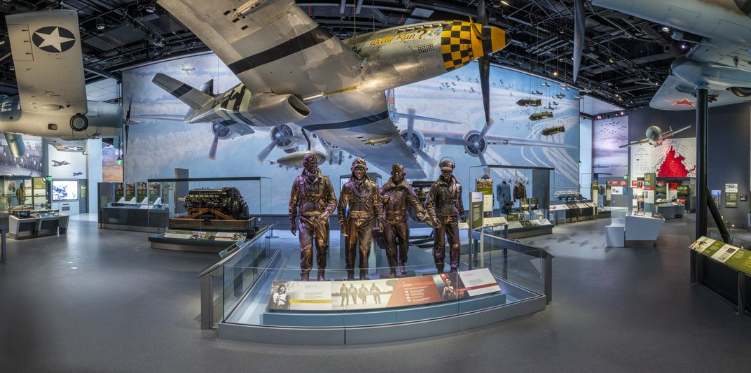 an exhibition gallery featuring hanging airplanes, four metal statues of soldiers and a large airplane illustration in the background