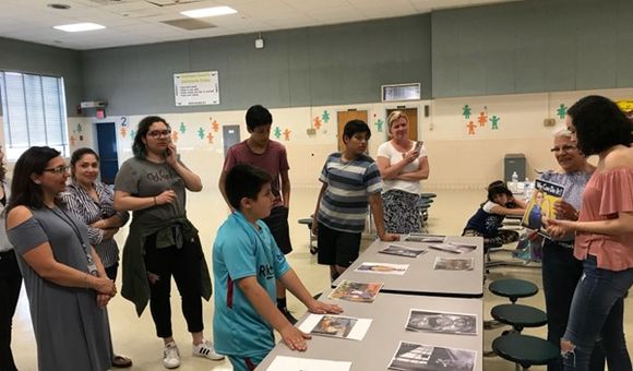 Group of adults and middle school children standing and actively discussing a set pf printed pictures laid out on a central table.