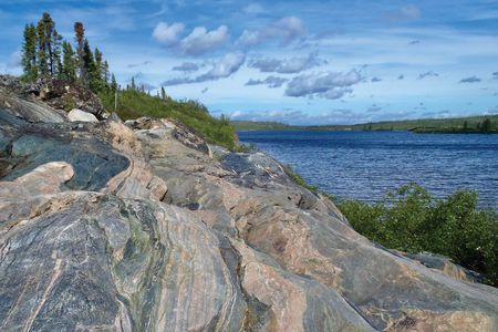 A slab of multicolored, sedimentary rock is pictured with vibrant trees and water surrounding it.