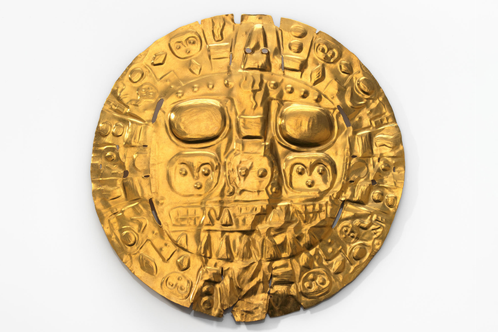 Against a white background, a shiny golden disc with raised features that depict two large, rounded eyes, the suggestion of a kind of face, and many interlocking diamonds, circles, lines and curved shapes