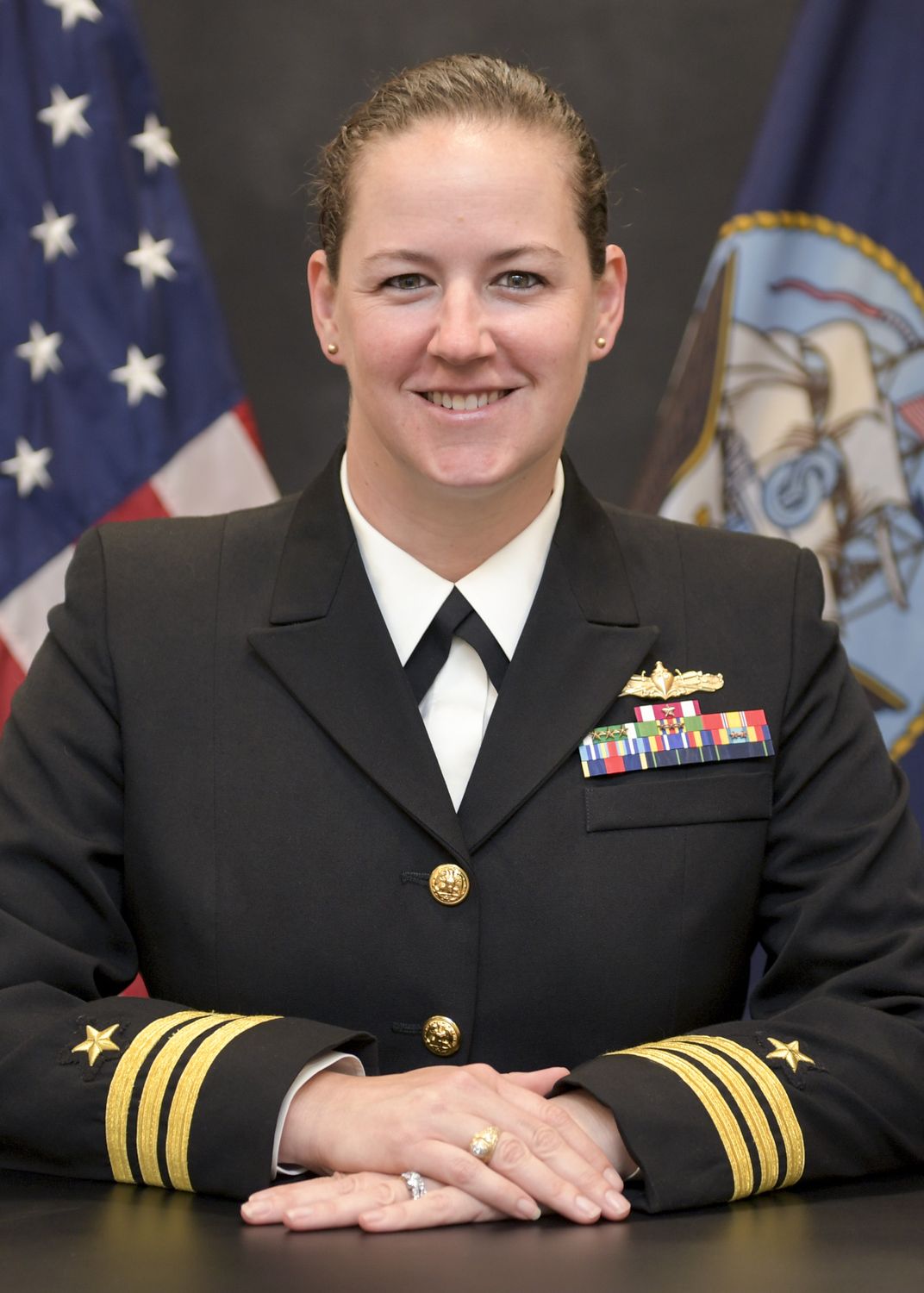 Young woman in black navy dress uniform smiling in front of the U.S. flag