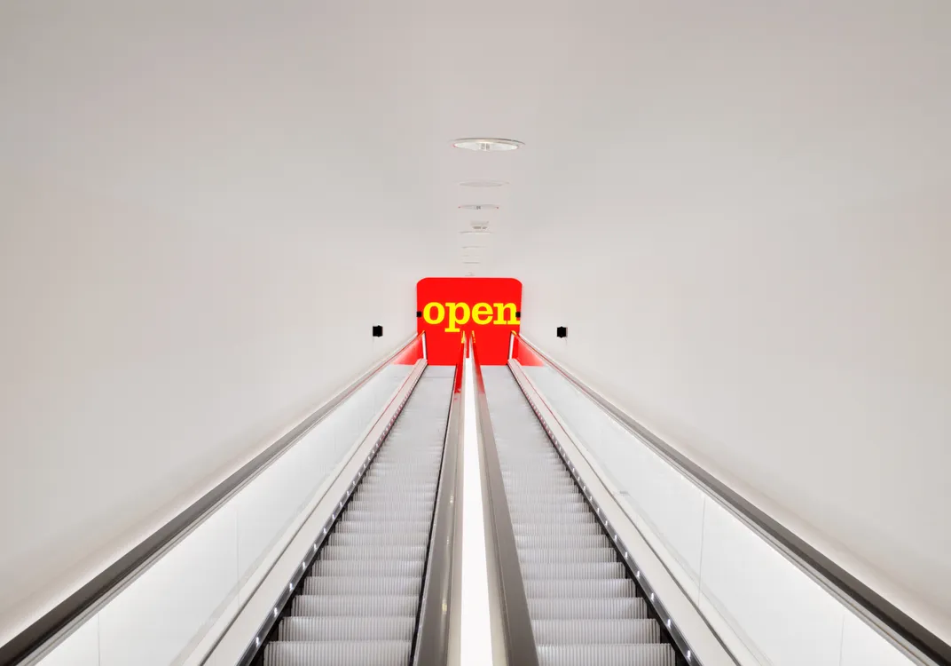 White escalator with glass sides leading to a red "open" sign in yellow letters, set in a minimal white interior.