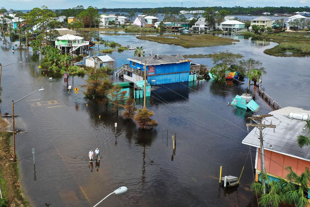 A flooded neighborhood from a bird's eye perspective, with two small figures walking in water that comes up to their calves. The street is entirely submerged, as are the foundations of the colorful houses