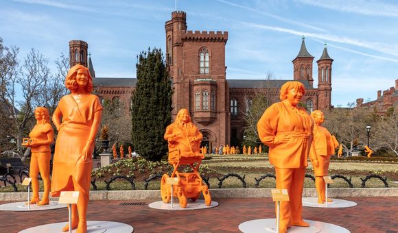 Five orange, 3D-printed statues of female scientists in a garden in front of the Smithsonian Castle on a sunny day.